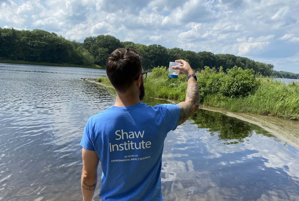 Dr. Charles Rolsky lifts a container of water to view it with his back facing the camera. He wear a light blue, Shaw Institute branded tee shirt and is facing towards a body of water with green trees surrounding the edges in the distance.