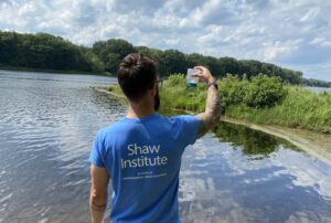 Dr. Charles Rolsky lifts a container of water to view it with his back facing the camera. He wear a light blue, Shaw Institute branded tee shirt and is facing towards a body of water with green trees surrounding the edges in the distance.