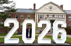 Hana stands inside the "0" of a large 2026 sign display in her blue cap and gown, in front of an brick, university building.