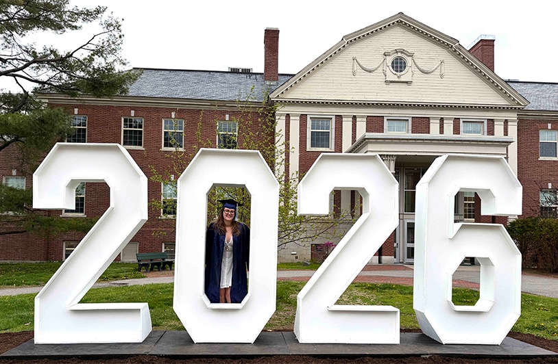 Hana stands inside the "0" of a large 2026 sign display in her blue cap and gown, in front of an brick, university building.