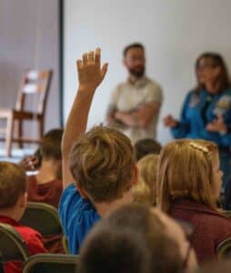 student raises hand to be called on for a question. He is the focus of the photo.