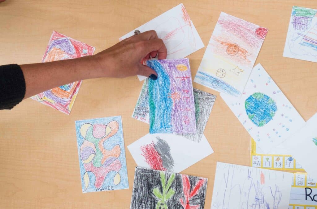 A hand reaches into the frame of the photo to pick up a hand colored postcard that will fly to space and then be returned to the student who drew it. All postcards are drawn with bright crayon colors on white paper the size of a standard postcard.