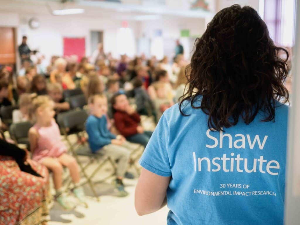 A woman with brown hair is framed in the image from the back, she's wearing a blue Shaw Institute logo tee shirt and looks out into a group of students that have assembled for a presentation.