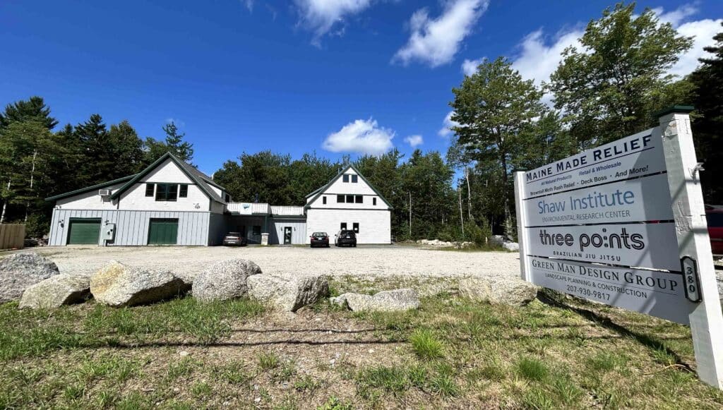 Shaw Institute's Environmental Research Center building with a green lawn and a deep blue sky overhead. A white wooden sign identifies the businesses inside the building.