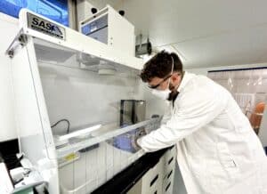 Dr. Charles Rolsky, in a white lab coat, with full PPE conducts research on a sample inside a fume hood in a white lab